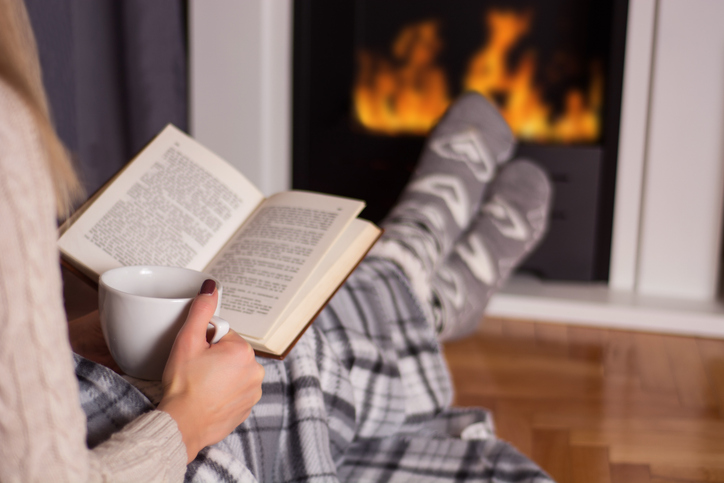 Beautiful young woman in front of the fireplace reading book and warming feet on fire and legs are covered with blanket, in hand holds cup of hot tea. Winter and cold weather concept at home. Close up, selective focus
